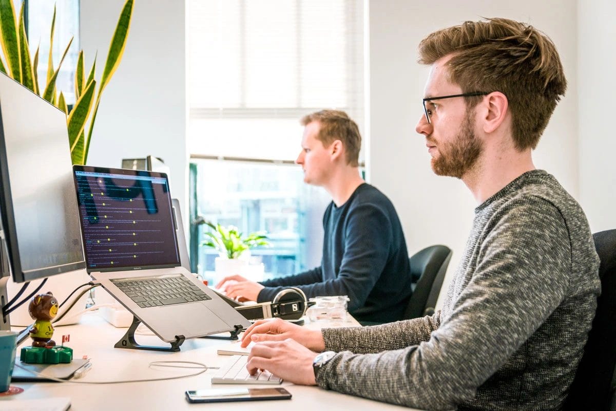 Two men working at desks with laptops in a bright office.